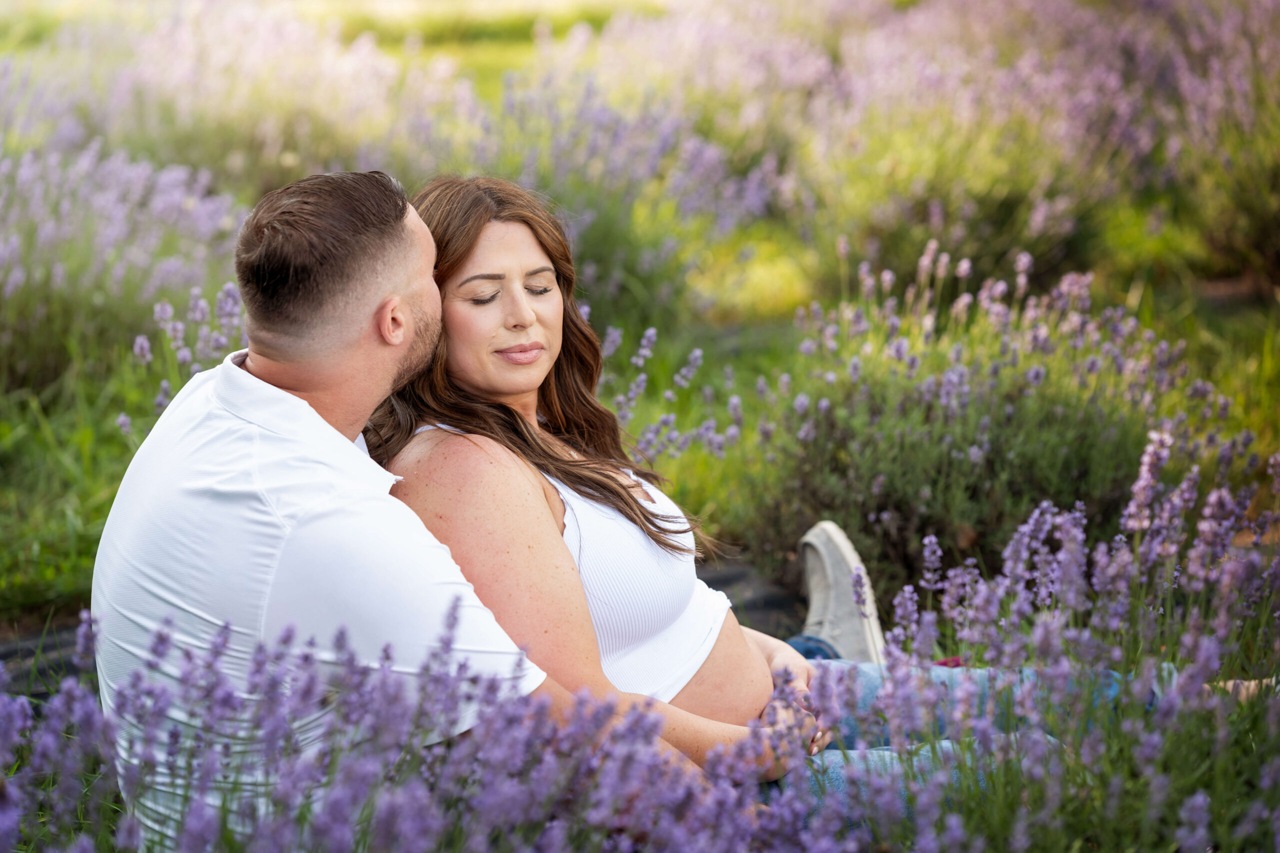 A happy pregnant couple snuggles in a field of lavender in jeans and white shirts after meeting with holistic beginnings
