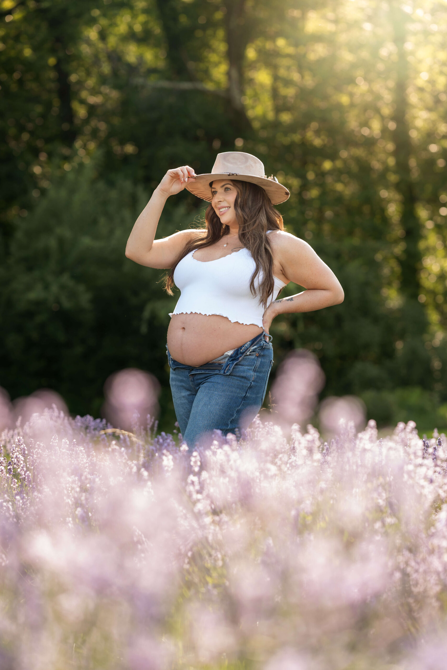 A mother to be stands with a hand on her hat brim with her bump out in a field of purple wildflowers in jeans and white blouse after visiting holistic beginnings