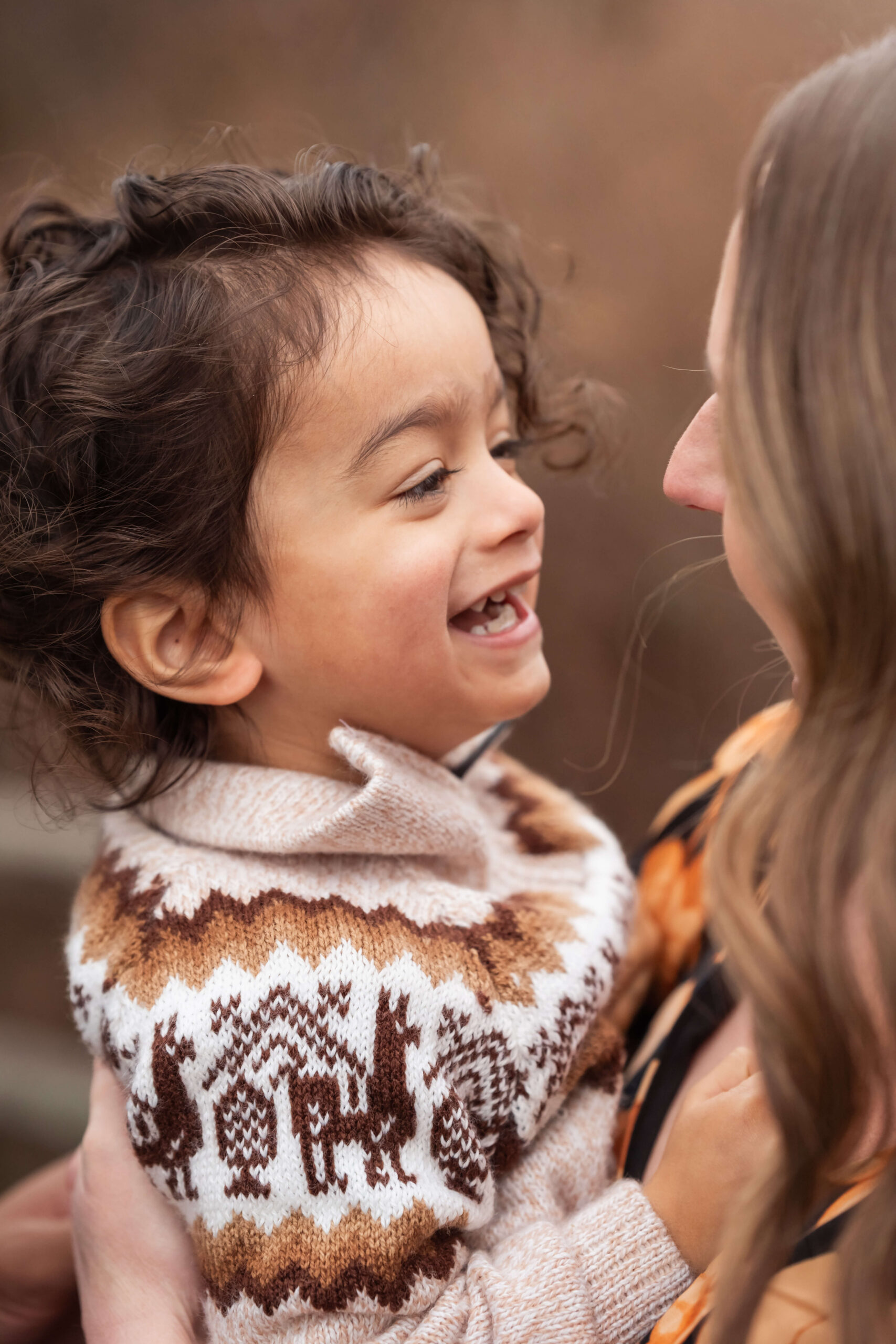 A giggling toddler in a knit brown and tan sweater in mom's arms
