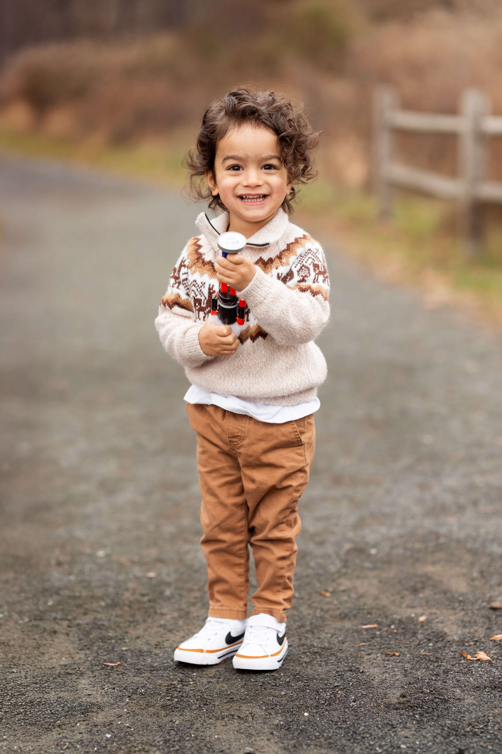A happy toddler smiles while playing wiht a toy in a park trail in a cream sweater