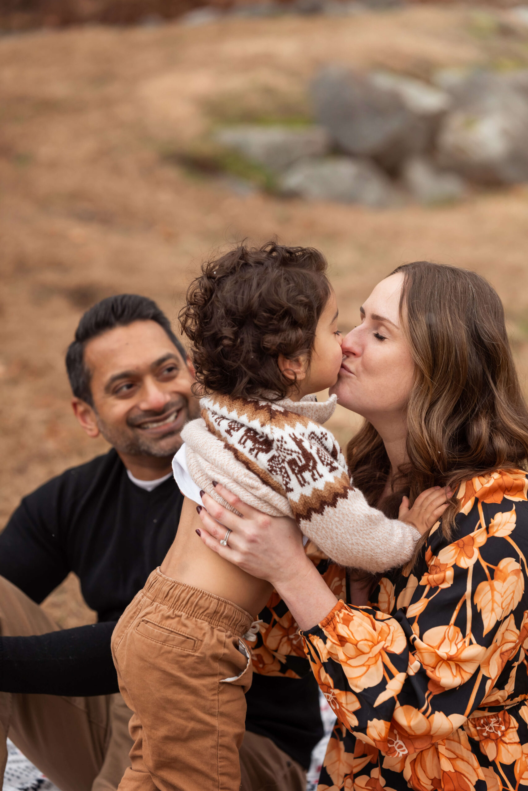 A happy mom kisses her toddler while sitting in a park with dad after some great mommy and me classes morris county nj
