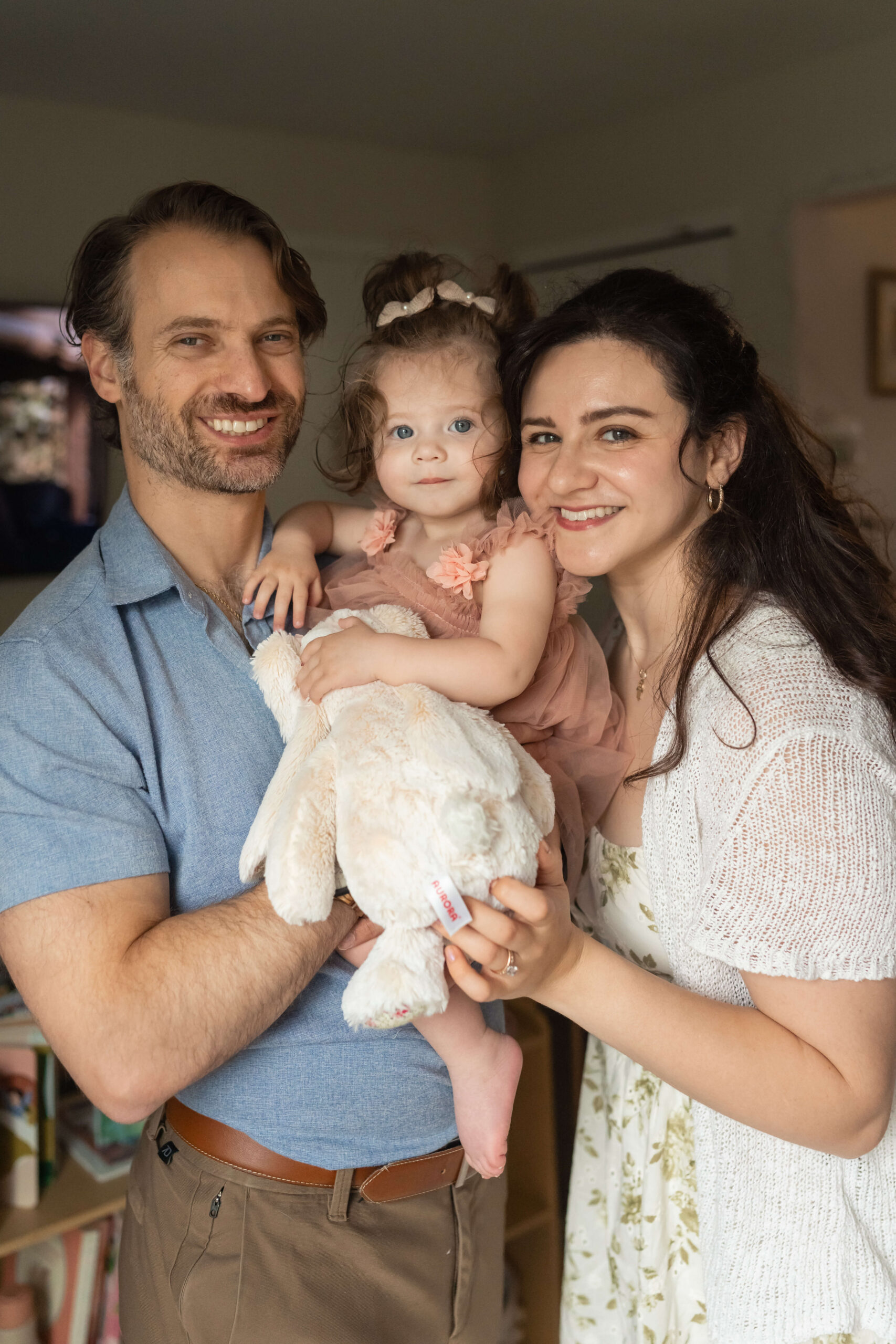 A mom and dad stand in a nursery holding their toddler daughter and her stuffed bunny with big smiles