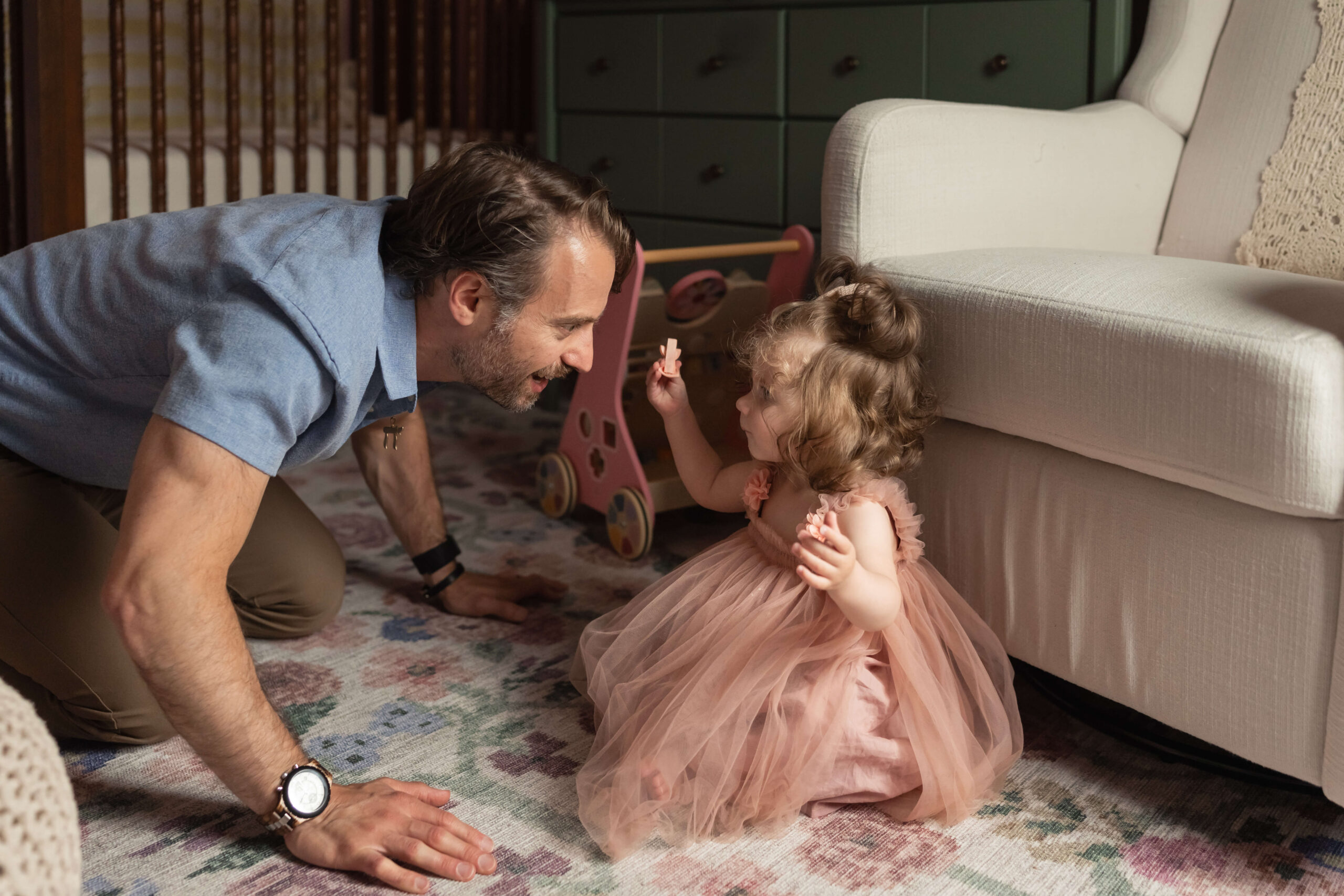 A happy toddler girl in a pink dress plays in the living room with dad on his hands and knees after some mommy and me classes in somerset county, NJ