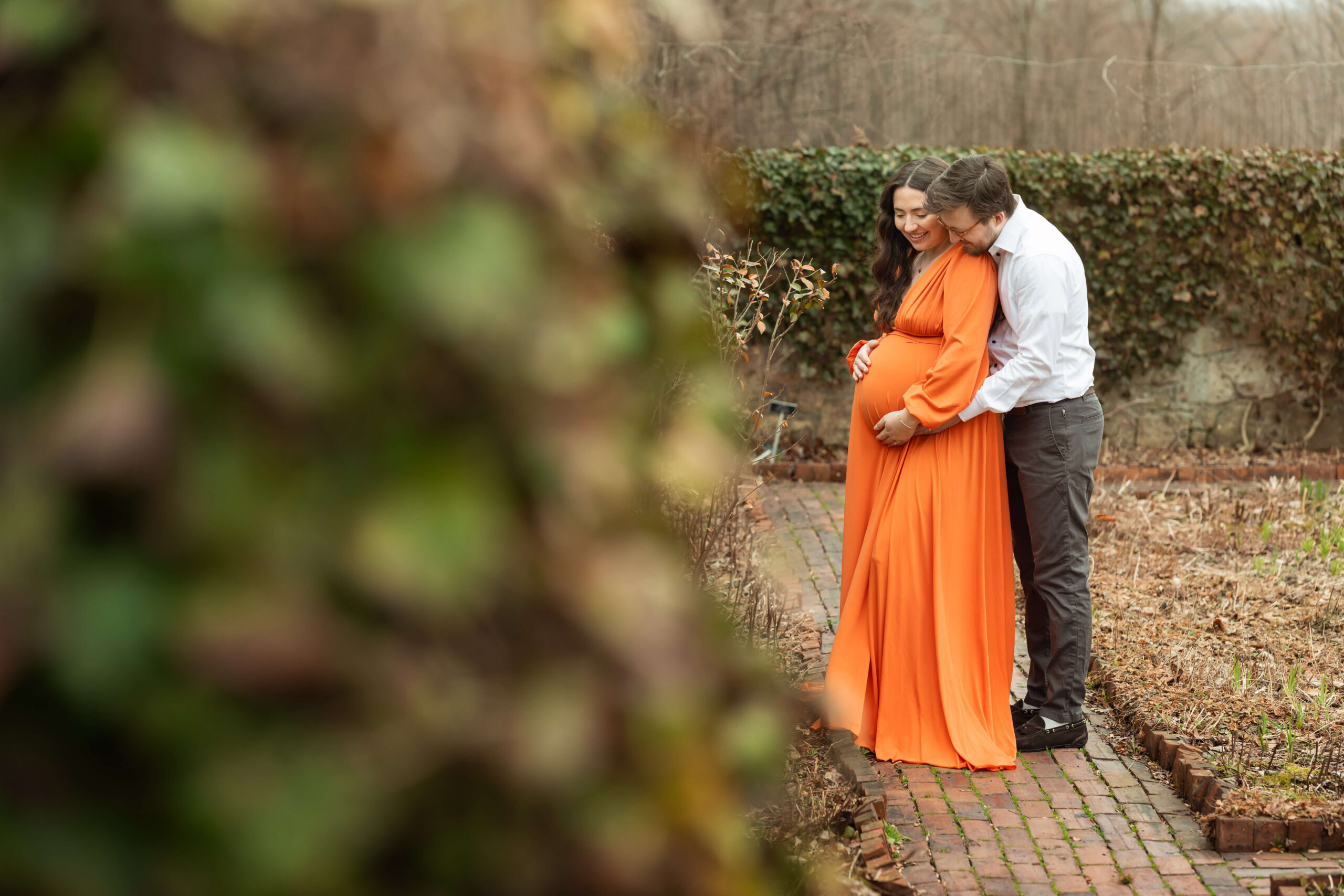 An expectant couple snuggles over the bump in an orange maternity gown while standing in a garden path after visiting morristown maternal fetal medicine