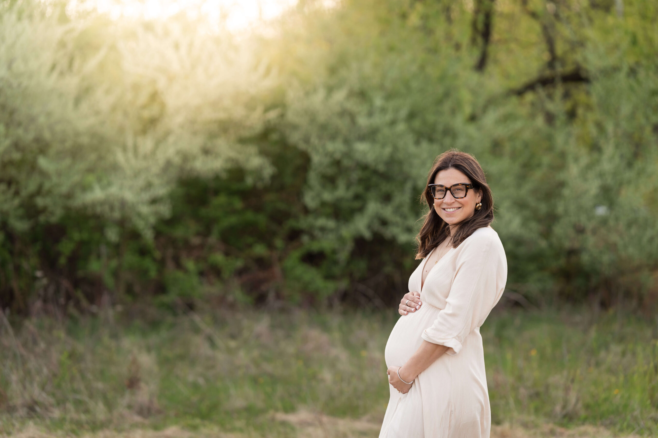 A happy mother to be in a white maternity dress stands in a park at sunset holding hands on her bump