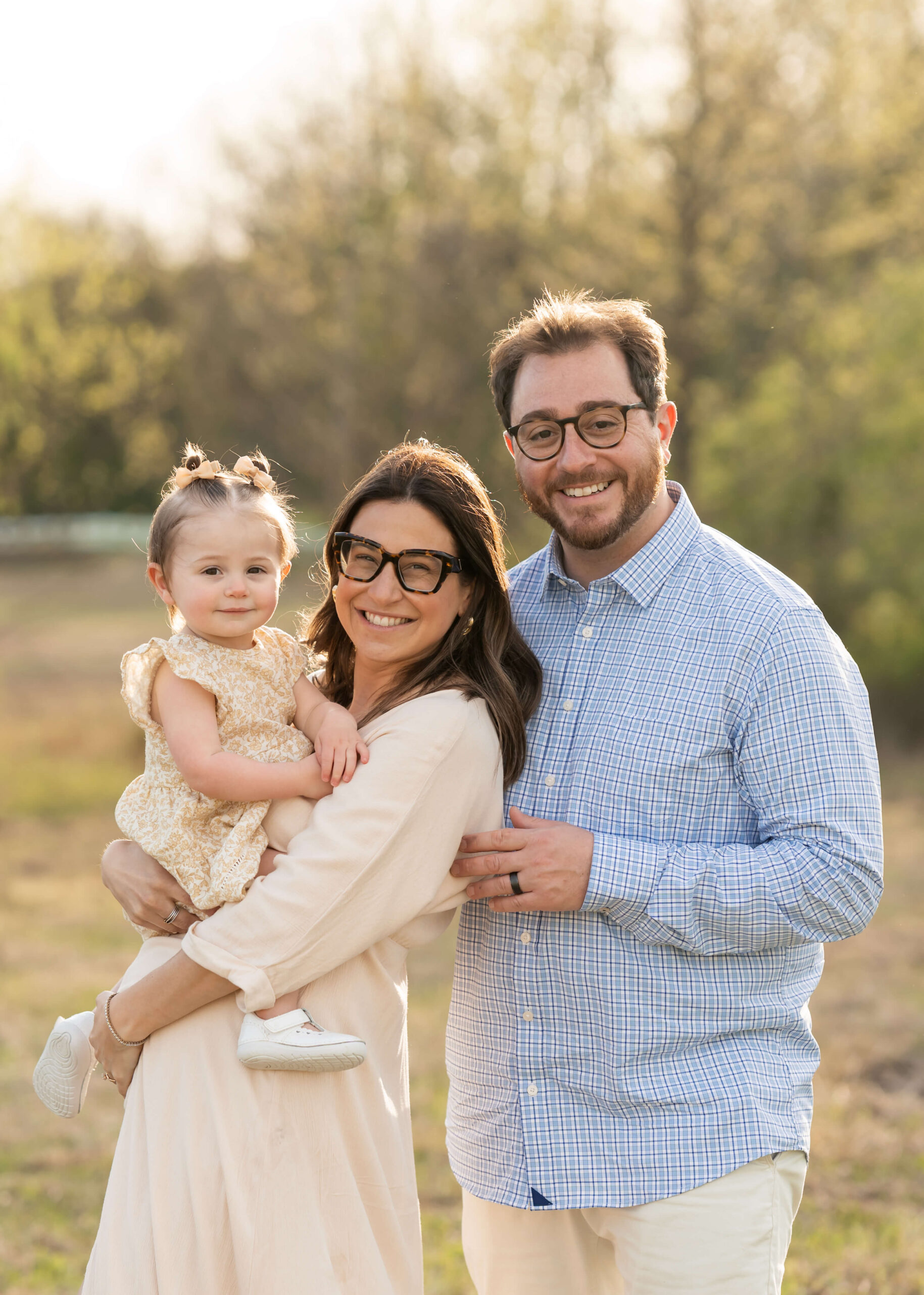 Expectant mom and dad stand in a park lawn at sunset holding their toddler daughter after visiting their obgyn morris county nj
