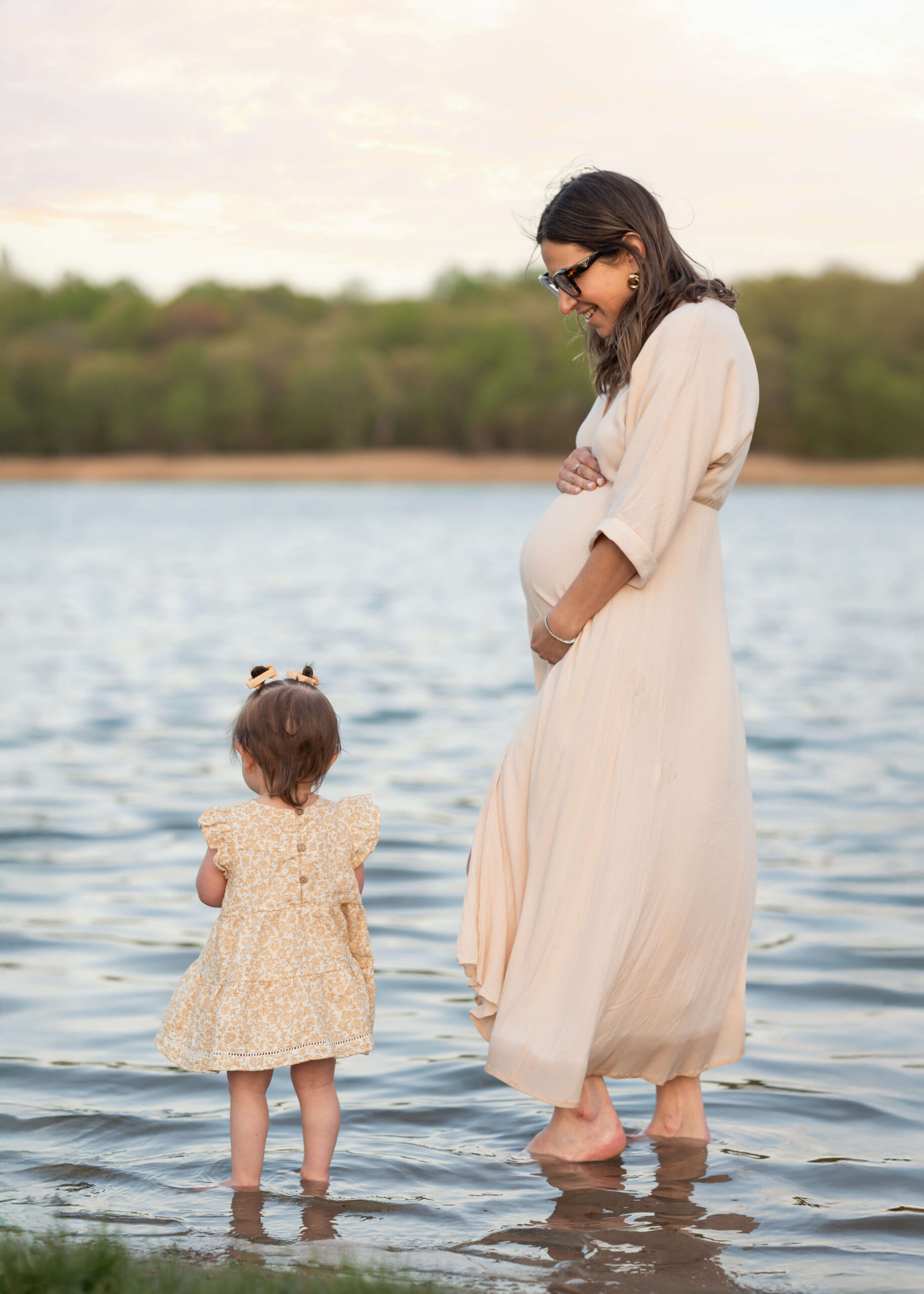 A pregnant mother in a white dress stands in a shallow lake with her toddler daughter at sunset thanks to an obgyn morris county nj