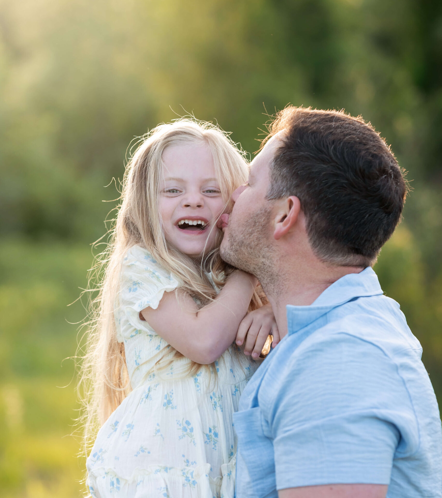 A happy dad kisses his giggling toddler daughter on the cheek in a white dress in a park at sunset