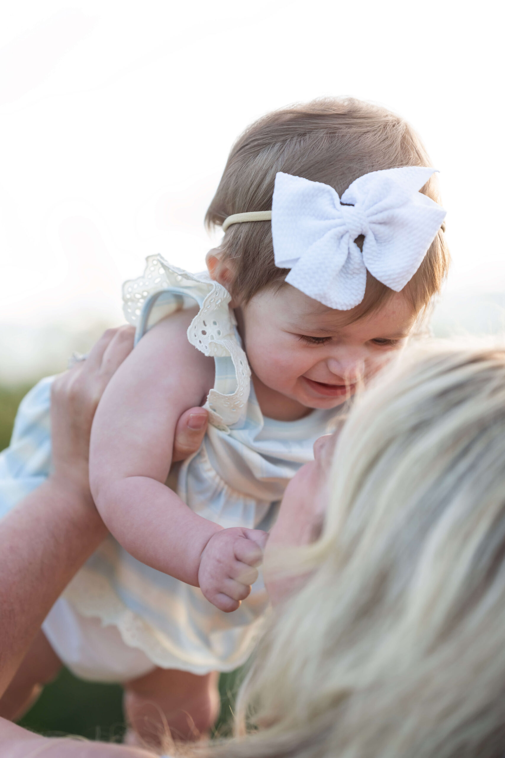 A toddler girl in a white dress is lifted by mom at sunset over her head