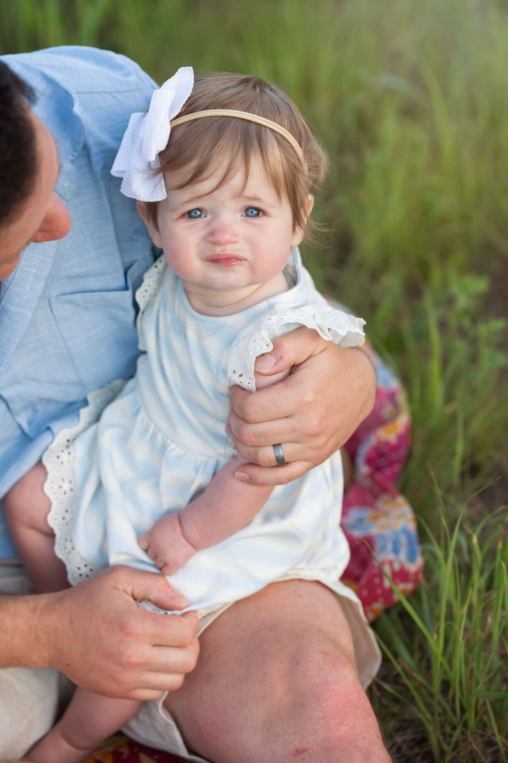 A toddler girl sits in dad's lap with a big bow and white dress in a park lawn thanks to postpartum place in chatham