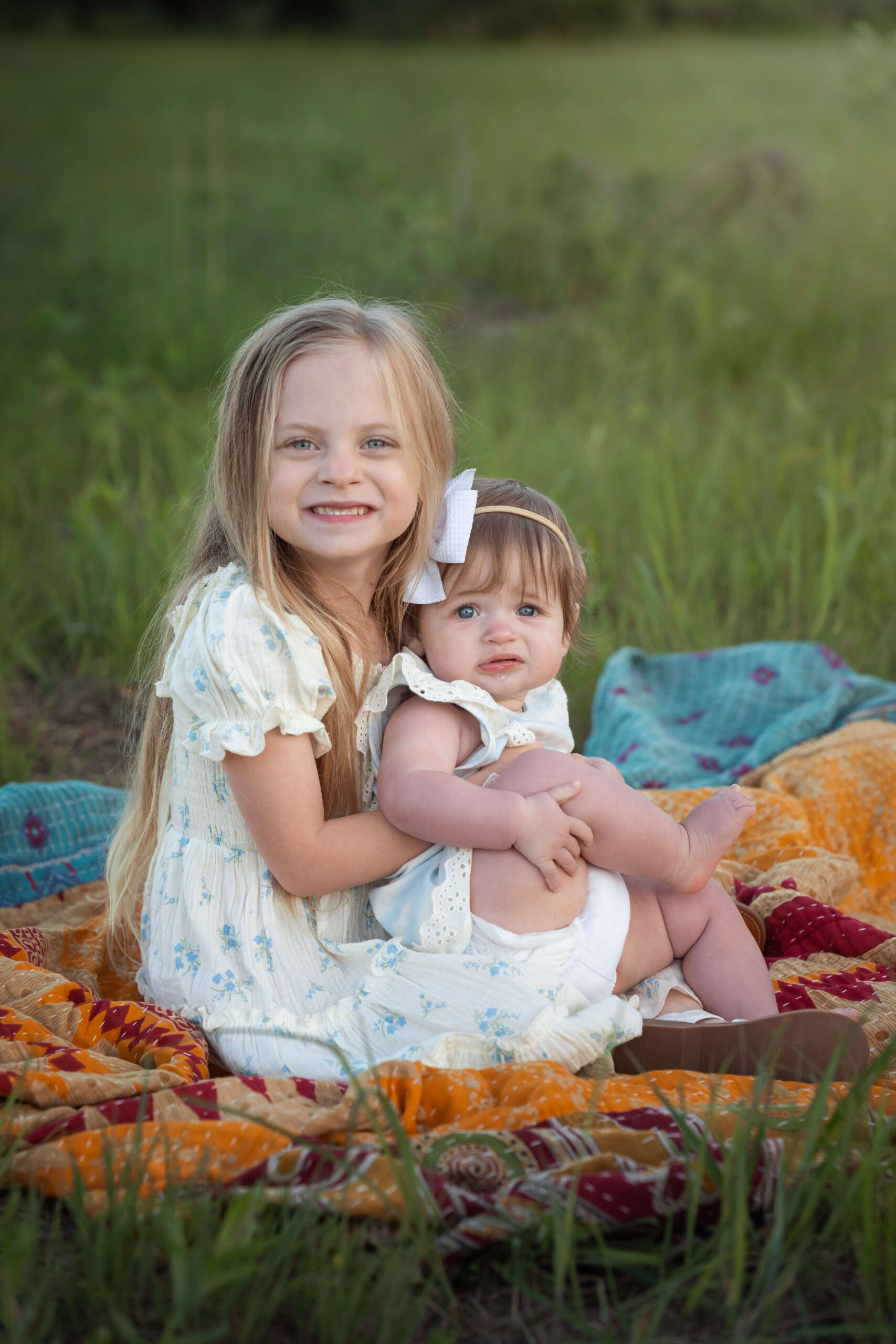 A baby girl sits in big sisters lap in white dresses on a picnic blanket in a lawn at sunset thanks to postpartum place in chatham