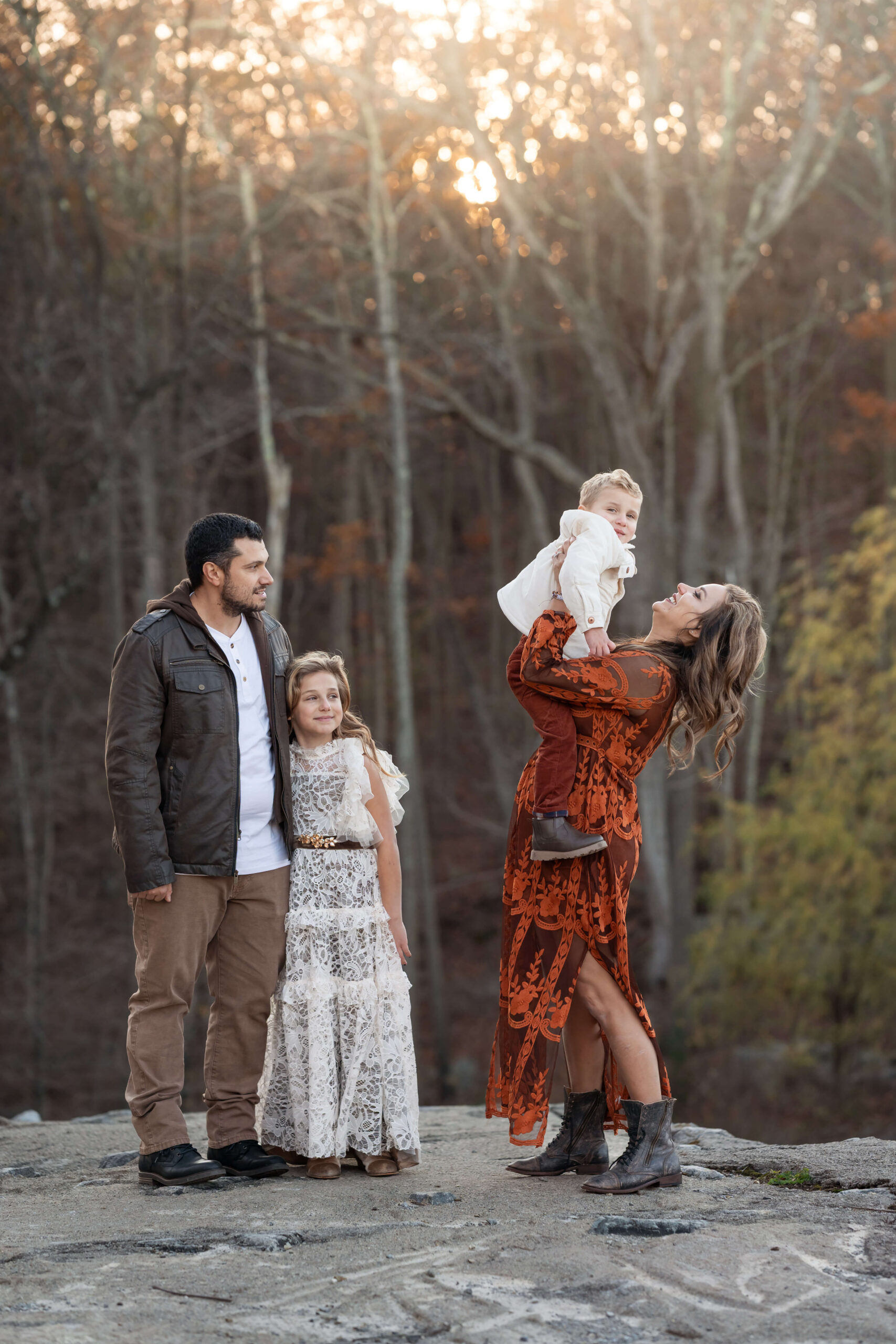 A happy mom in a rust lace gown lifts her toddler while standing on a boulder at sunset with her husband and daughter watching