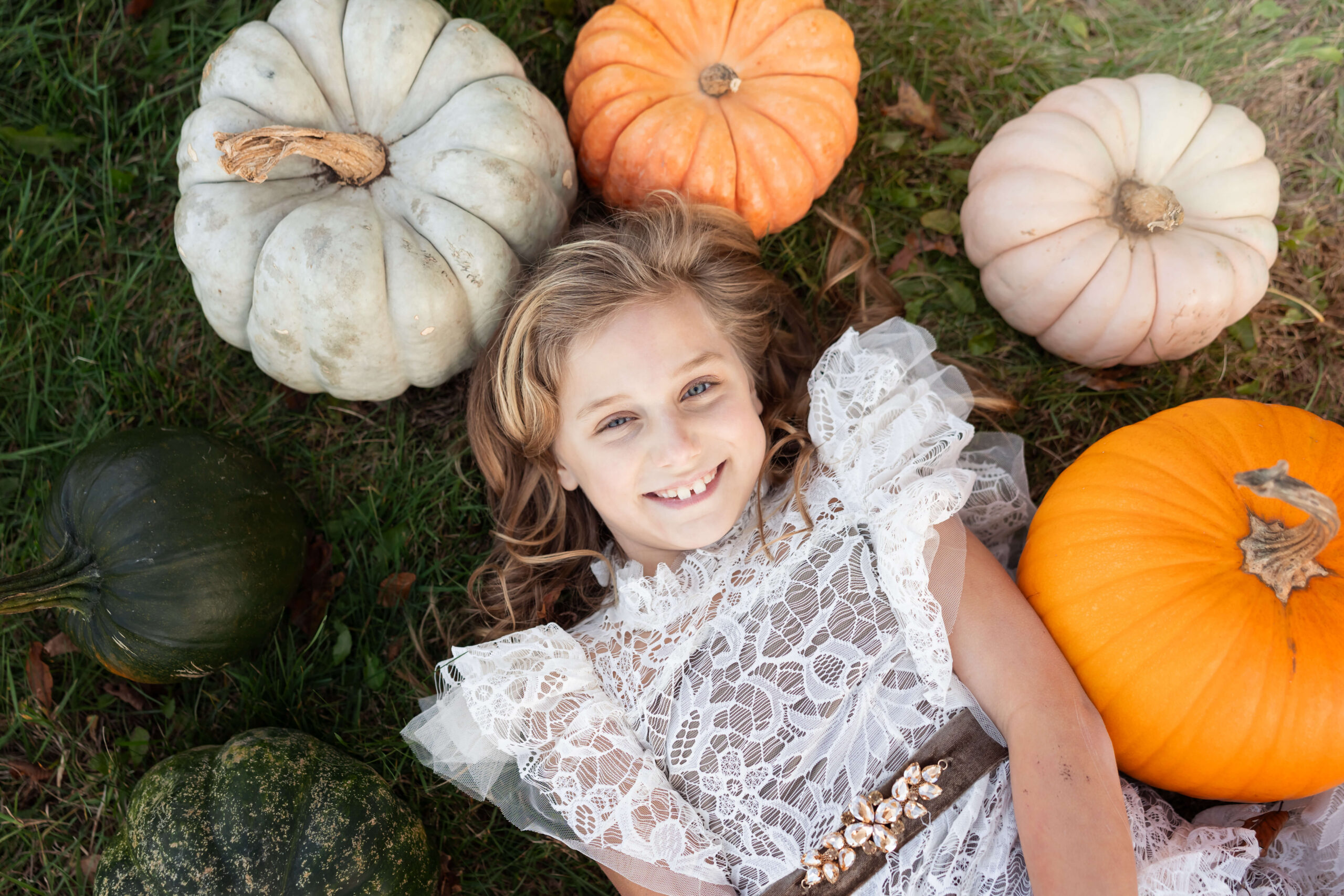 A toddler girl in a white lace dress lays surrounded by pumpkins in the grass after visiting wildflowers boutique