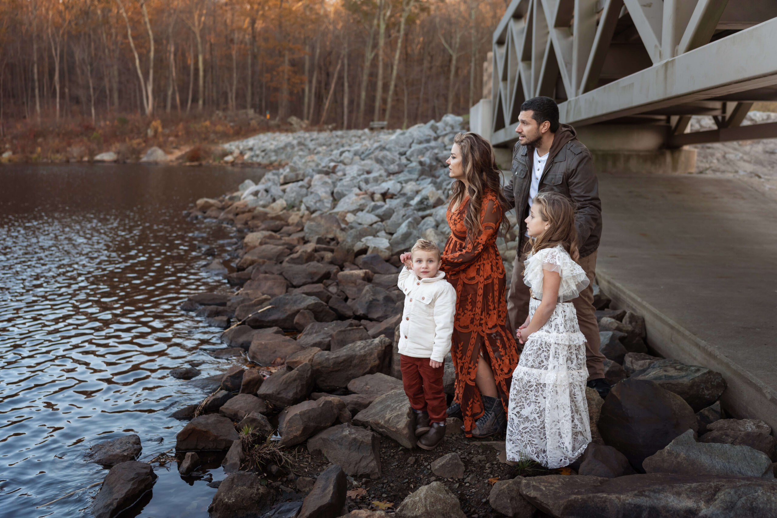 A mom and dad stand on rocks under a bridge on a lake with their young son and daughter holding hands after exploring wildflowers boutique